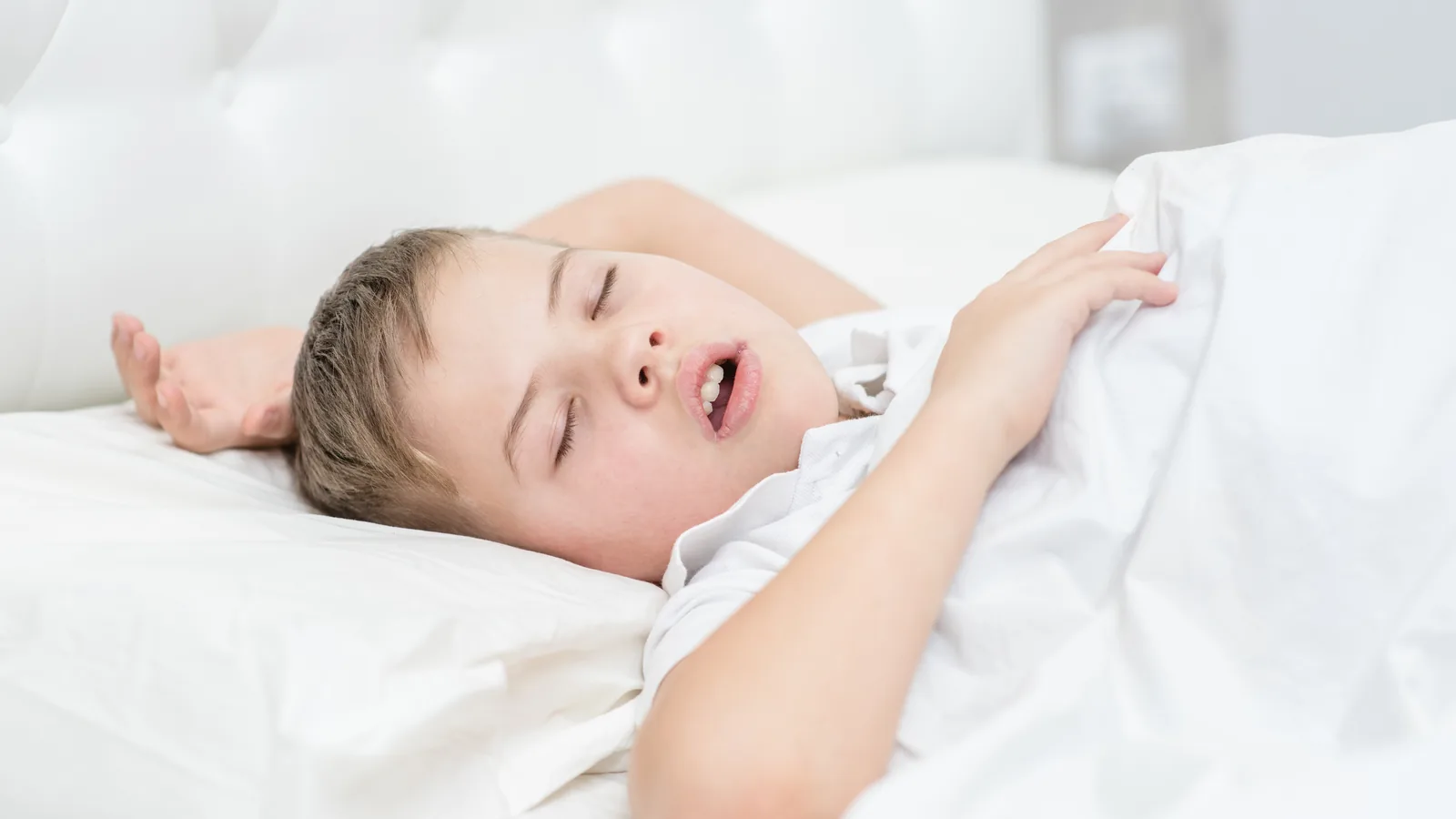 Criança dormindo de boca aberta na cama, com a cabeça inclinada para trás — o padrão de quem respira pela boca durante o sono.