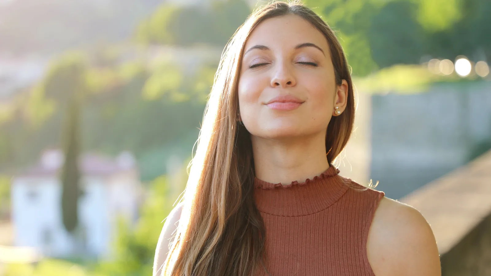 Mulher de olhos fechados respirando fundo pelo nariz ao ar livre, com vegetação e luz do fim de tarde ao fundo.