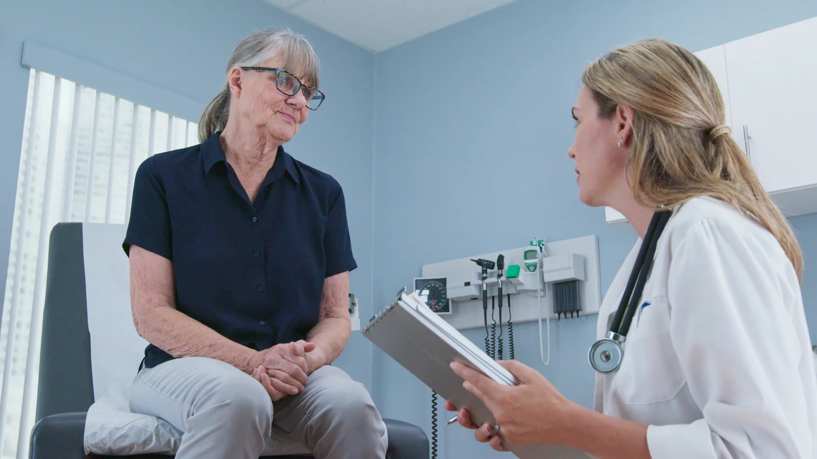 Médica de jaleco com prancheta na mão conversando com uma paciente idosa sentada na maca do consultório, colhendo a história clínica.
